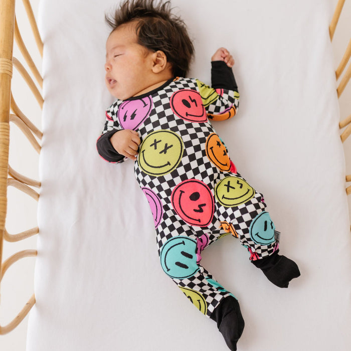 Baby wearing a colorful smiley face onesie lying on a white blanket.