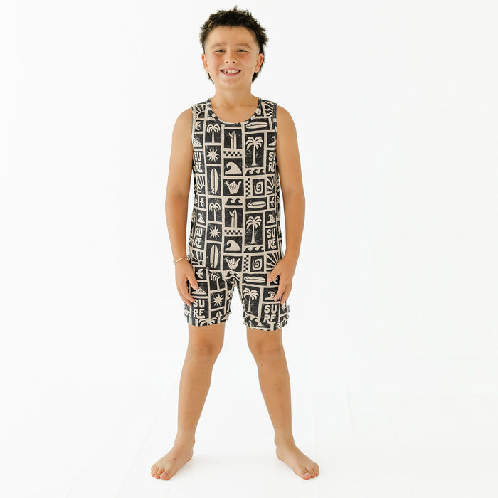 A young boy stands barefoot against a plain white studio background, wearing a sleeveless black and white patterned top and matching shorts.