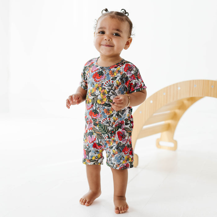 Child wearing a colorful floral romper standing on a white floor with a wooden toy in the background.