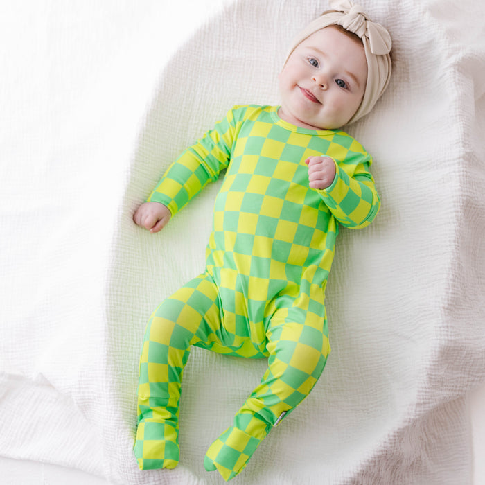 Baby wearing a green and yellow checkered outfit lying on a white blanket.