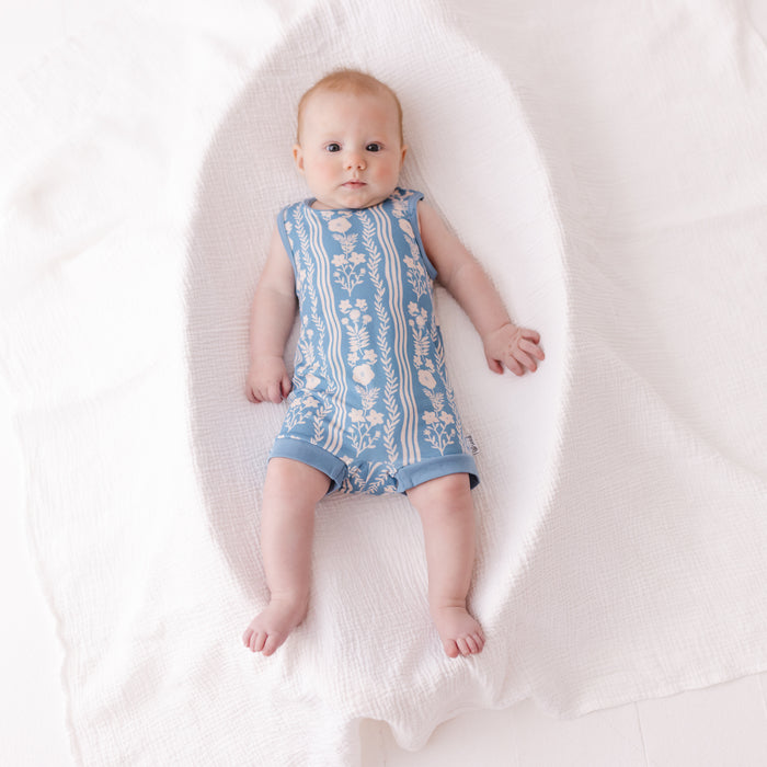 A baby lying on a white textured blanket, wearing a sleeveless light blue romper with a white floral pattern.